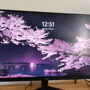 Könnte beinhalten: Ein schwarzer LG-Computermonitor, der ein digitales Bild eines Kirschblütenbaums mit rosa Blüten anzeigt. Der Bildschirm zeigt die Uhrzeit 12:51 und das Datum, Dienstag, 3. Oktober. Der Monitor steht auf einem Holztisch.