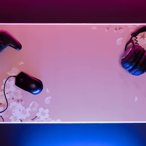 May include: A pink gaming mouse pad with a floral design, a black wireless mouse, a black video game controller, and black over-ear headphones.