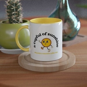 May include: A white ceramic mug with a yellow rim and handle. The mug has a graphic of a smiling sun with the text "a cupful of sunshine" printed on it.