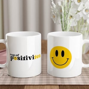 May include: Two white ceramic mugs with a black and yellow design. One mug has a smiley face and the other says "cup of positivitea" with a smiley face.
