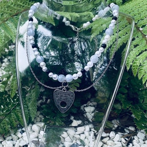 May include: A silver necklace with a heart-shaped pendant featuring a Ouija board design. The necklace is layered with two strands of beads, one strand of white and blue beads and another strand of black and white beads.