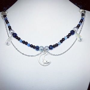 May include: A silver chain necklace with a crescent moon charm and a beaded strand of black and blue beads. The necklace features small star and teardrop charms.