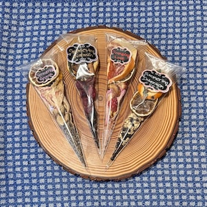 May include: Four clear plastic cones filled with dried fruit and flowers. Each cone has a black chalkboard tag with white text that reads "Simmering Growth", "Simmering Warmth", "Simmering Love", and "Simmering Intention". The cones are arranged on a wooden slice.