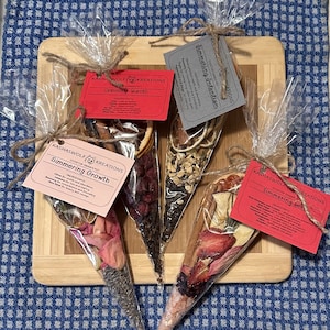 May include: Four clear plastic cones filled with dried herbs, flowers, and spices. Each cone has a red label with the text "KASHASWOLF KREATIONS" and the name of a simmering blend: "Simmering Intention", "Simmering Warmth", "Simmering Growth", and "Simmering Love".