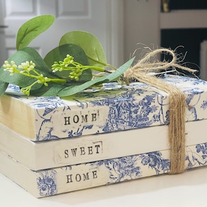 May include: A stack of decorative books featuring a blue and white floral design, with the words "HOME SWEET HOME." The books are tied with twine and decorated with artificial greenery. Ideal for home decoration.