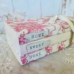 May include: A stack of decorative books featuring a pink floral toile design. Each book has a label with the words "HOME SWEET HOME". The books are tied with twine and a lace ribbon, evoking a vintage style.