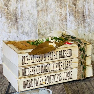 May include: A stack of three decorative wooden books with white painted sides and tops. The books have brown text that reads "ELF-KISSED & FAIRY BLESSED", "WHISPER OF MAGIC IN EVERY LEAF", and "WHERE FAIRY FOOTPRINTS LINGER". The books are decorated with a feather, flowers, and twine.