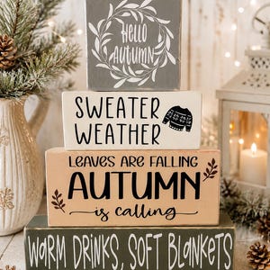 May include: A stack of four rectangular wooden blocks with painted text and graphics. The top grey block says Hello Autumn. The white block says Sweater Weather with a sweater icon. The tan block says Leaves Are Falling Autumn is calling. The bottom green block says Warm Drinks, Soft Blankets.