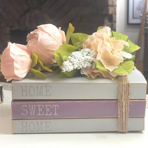 May include: A decorative stack of three wooden books with the words "HOME SWEET HOME" in white lettering. The books are painted in light grey and lavender, and are adorned with faux peach-coloured peonies and other flowers. A twine bow adds a rustic touch.