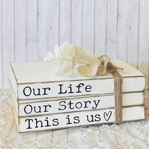 May include: Three white wooden blocks, tied with twine, are stacked together. Each block has a phrase printed on it: "Our Life", "Our Story", and "This is us". A sprig of dried flowers sits on top.