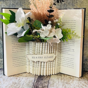 May include: A decorative book sculpture with a floral display. The open book is adorned with white and green flowers, dried grasses, and a tag reading "MR & MRS SLOANE". The pages are folded to form a vase-like structure.