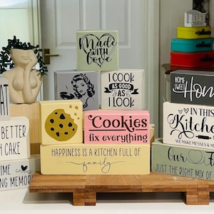May include: A collection of wooden signs with various kitchen-themed sayings, including "Let's Eat", "Life is Better with Cake", "Cookies Fix Everything", "Happiness is a Kitchen Full of Family", "Home Sweet Home", "In This Kitchen We Make Messes & Memories", and "Our Family Just the Right Mix of Chaos & Love".