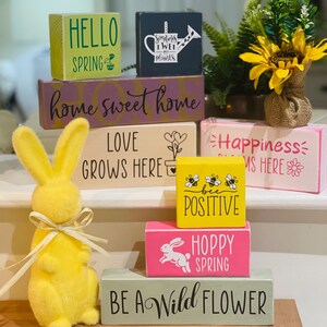 May include: A stack of wooden blocks with spring-themed sayings, including "Hello Spring", "Home Sweet Home", "Love Grows Here", "Happiness Blooms Here", "Be Positive", "Hoppy Spring", and "Be A Wild Flower". A yellow bunny figurine sits in front of the blocks.