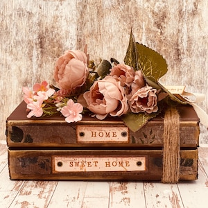 May include: Two decorative brown boxes stacked, with labels reading "HOME" and "SWEET HOME". The boxes are decorated with pink flowers and greenery, tied with twine and a ribbon. The background is a light brown, wood-effect surface.