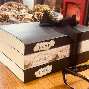 May include: A stack of decorative books painted black, tied with a black ribbon. The books have the words "HOME SWEET HOME" on them. The middle book has a floral design. A pair of black-framed glasses rests on the side.