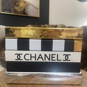 May include: A collection of decorative books in a stack. The top book is gold, followed by a black and white striped book, a white book with the word "CHANEL" in black, and a black book. The bottom book is silver.