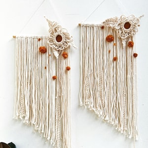 Op de afbeelding: Twee macramé wandkleden in een natuurlijke, gebroken witte kleur. Elk heeft een zonnebloemontwerp en kleine oranje accenten. De wandkleden zijn opgehangen aan houten stokken en hebben een gefranjerde onderkant, wat een bohemien esthetiek creëert.