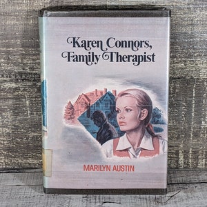 May include: A vintage book titled "Karen Connors, Family Therapist" by Marilyn Austin. The cover features an illustration of a woman with blonde hair, a house, and a silhouette of a man. The book has a light-colored background and is set against a wooden surface.
