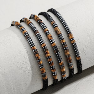 Könnte beinhalten: Fünf Armbänder mit Perlen und einem schwarzen Band. Die Armbänder haben eine Kombination aus braunen, silbernen und schwarzen Perlen. Die Perlen sind in einem sich wiederholenden Muster angeordnet.