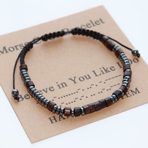 May include: A black cord bracelet with brown wooden beads and black hematite beads. The bracelet has a sliding knot closure. The bracelet is on a brown card with the text "Morse Bracelet Believe in You Like I Do".