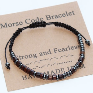 Può includere: Un braccialetto con codice Morse con perline di legno marrone scuro e perline di metallo color argento su un cordoncino nero. Il braccialetto è esposto su una carta marrone con il testo "Morse Code Bracelet" e "Strong and Fearless".