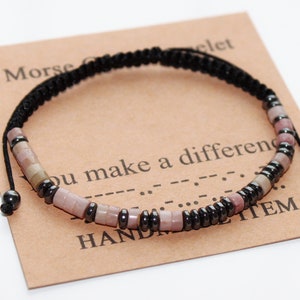 May include: A black cord bracelet with pink and grey beads and black metal beads. The bracelet is on a brown card with the text "Morse Code Bracelet" and "You make a difference" in black text.