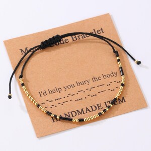 Peut inclure: Un bracelet noir réglable avec des perles dorées et des perles cylindriques noires. Le bracelet est présenté sur une carte marron avec le texte "Morse Code Bracelet" et "I'd help you bury the body". La carte indique également "HANDMADE ITEM".