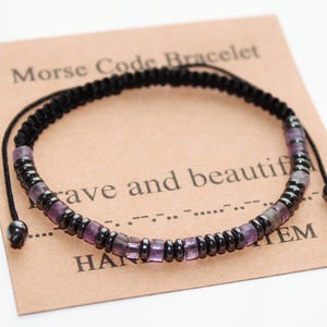 May include: A black Morse code bracelet with purple and black beads, presented on a brown card. The card says "Morse Code Bracelet" and "brave and beautiful". The bracelet is handmade.
