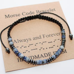 Könnte beinhalten: Ein geflochtenes Morsecode-Armband in Schwarz mit blauen und grauen Perlen. Das Armband liegt auf einer braunen Karte mit den Aufschriften "Morse Code Bracelet" und "Always and Forever". Auf der Karte steht auch "HANDMADE ITEM".