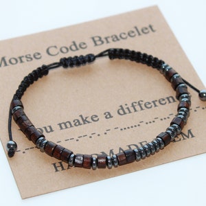 Puede incluir: Un brazalete de cordón negro con cuentas de madera marrón y cuentas de metal plateado. El brazalete está sobre una tarjeta marrón con el texto "Morse Code Bracelet" y "You make a difference" en código Morse.