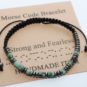 Può includere: Un braccialetto con cordino nero con chiusura a nodo scorrevole. Il braccialetto presenta una combinazione di perline grigio scuro e turchese, disposte a motivo. Il braccialetto è esposto su una carta marrone con il testo "Morse Code Bracelet" e "Strong and Fearless".