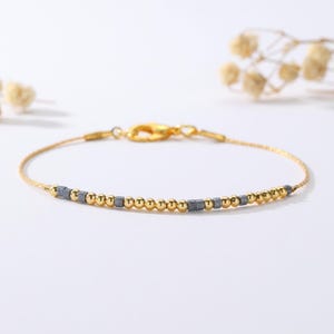 May include: A fine gold bracelet with a series of small, round gold beads and square grey beads. The bracelet has a simple design with a clasp closure. The jewelry is displayed on a white surface.