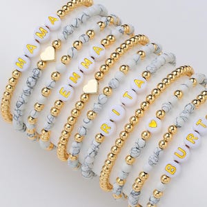 Könnte beinhalten: Eine Sammlung von Armbändern mit Perlen. Die Armbänder haben weiße und graue marmorierte Perlen, goldfarbene Perlen und weiße Buchstabenperlen, die Namen buchstabieren. Einige Armbänder enthalten goldfarbene herzförmige Charms.