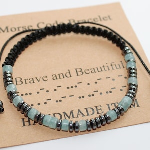 Op de afbeelding: Een handgemaakte Morsecode armband met een zwart geweven koord en verstelbare sluiting. De armband heeft lichtblauwe en donkergrijze kralen. De kaart leest "Brave and Beautiful" en "HANDMADE ITEM".