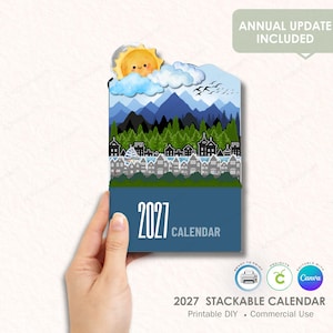 May include: A 2027 calendar featuring a layered illustration of a yellow sun, white clouds, blue mountains, a green forest, and a town with grey buildings. The text 2027 CALENDAR is printed in white on a dark blue background at the bottom.