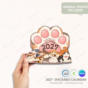May include: A 2027 calendar shaped like a large cat paw with pink pads. The design features a group of illustrated cats in various colours and poses resting at the base. The text reads: Sometimes I'm a... CALENDAR 2027. The calendar is held in a hand.