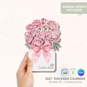 Könnte beinhalten: Ein stapelbarer Kalender 2027 mit floralem Design. Der Kalender zeigt einen Strauß rosa Pfingstrosen, eine rosa Schleife und den Text "2027 Calendar". Das Design enthält den Text "Annual Update Included" und Symbole für Druck und Bearbeitung.