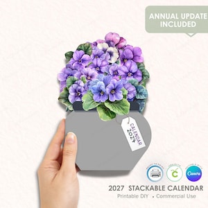 Puede incluir: Un calendario apilable 2027 en forma de corazón gris, con una ilustración de acuarela de flores moradas. El calendario incluye el texto "Annual Update Included" y está etiquetado como "Printable DIY • Commercial Use."