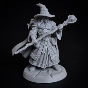 Può includere: Una miniatura in resina grigia raffigurante un mago con cappello a punta, una lunga veste con motivi complessi e un mantello. Il personaggio tiene in mano un bastone sormontato da una sfera e poggia su una base circolare effetto pietra.