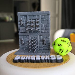 Peut inclure: Un mur de donjon gris imprimé en 3D avec des pointes et une fenêtre à barreaux, à côté d'un dé vert fluo à 20 faces. La base porte le texte "DUNGEON3D" en lettres blanches sur une base noire. Le mur est fait de briques grises.