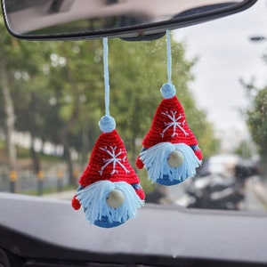 May include: Two red and white crocheted gnome ornaments with white beards and snowflakes hanging from the rearview mirror of a car.