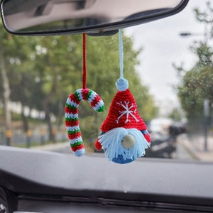 May include: A red and white crocheted candy cane and a red and white crocheted gnome with a blue beard hanging from the rearview mirror of a car.