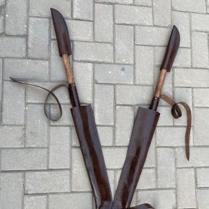 May include: Two brown leather sheathed knives with wooden handles are displayed on a gray brick surface. The knives are positioned in a V-shape, with leather straps and a belt. The sheaths are dark brown.