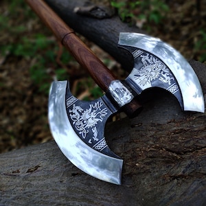 May include: A double-bladed axe with a dark wooden handle and silver blades. The blades feature intricate engraved designs, including a dragon motif. The axe rests on a weathered log, with a blurred green background.