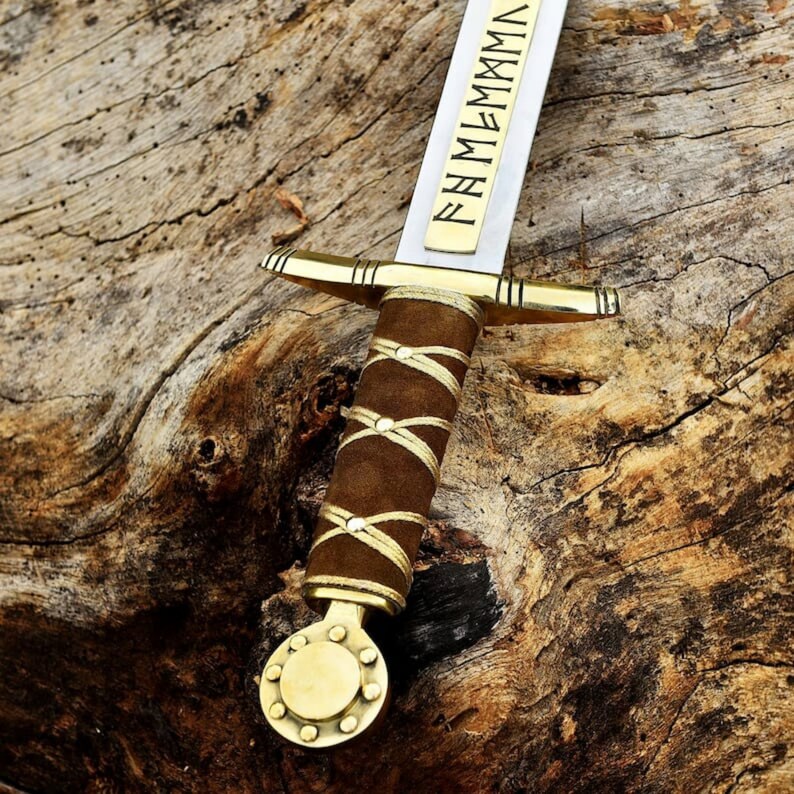 May include: A close-up of a decorative sword with a brown leather-wrapped handle and gold accents. The blade is silver with gold lettering. The sword rests on a textured wooden surface.