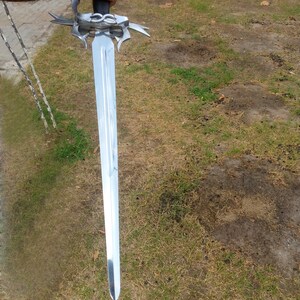 May include: A long, silver sword with a decorative hilt and a black handle. The blade is polished and reflects light. The sword is held against a background of green grass.