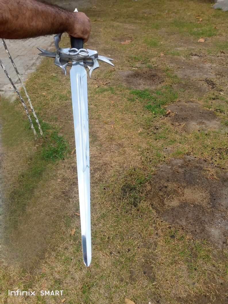 May include: A long, silver sword with a decorative hilt and a black handle. The blade is polished and reflects light. The sword is held against a background of green grass.