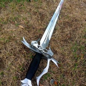 May include: A silver dagger with a double-edged blade, ornate guard, and a black wrapped handle. The dagger is lying on a bed of dry grass and some green grass.