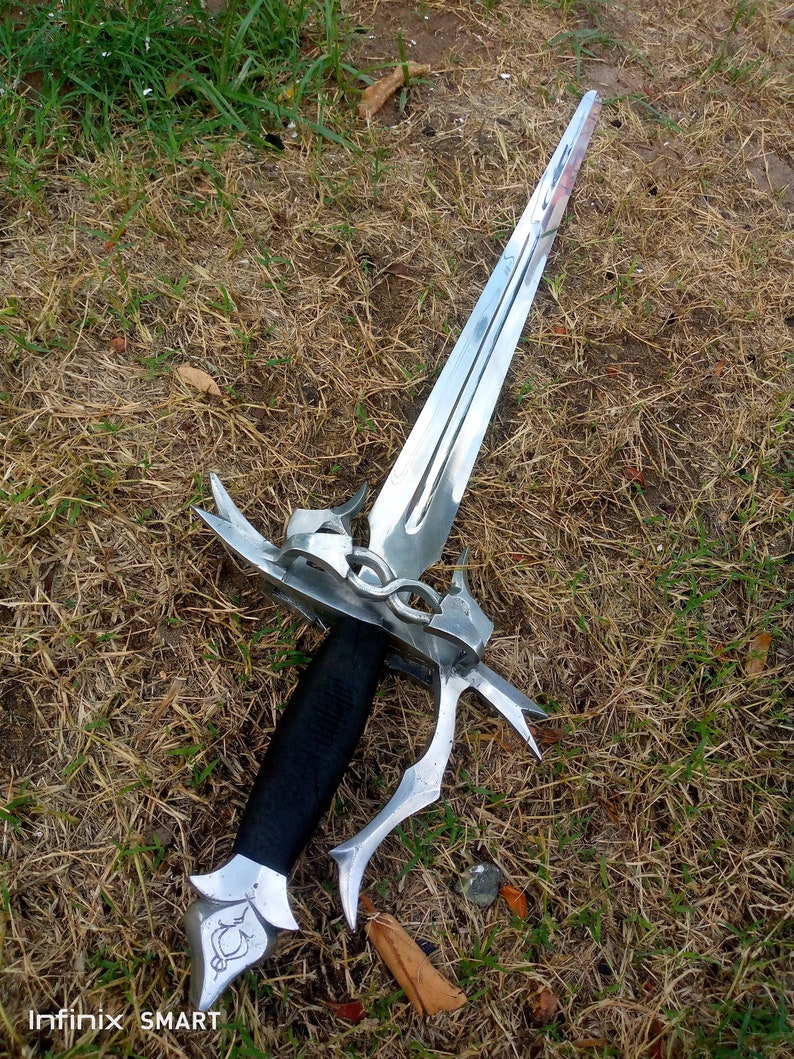 May include: A silver dagger with a double-edged blade, ornate guard, and a black wrapped handle. The dagger is lying on a bed of dry grass and some green grass.
