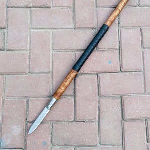 May include: A spear with a wooden shaft and a sharp, silver-colored blade. The shaft has a dark, wrapped grip. The spear is lying on a brick surface.